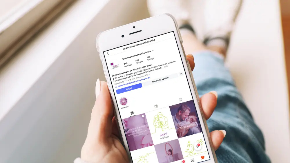 Eine Hand hält ein Smartphone, auf dem das Instagram-Profil des Kinderwunschzentrums Karlsruhe mit verschiedenen Beiträgen zu den Themen Kinderwunsch und Familie geöffnet ist.