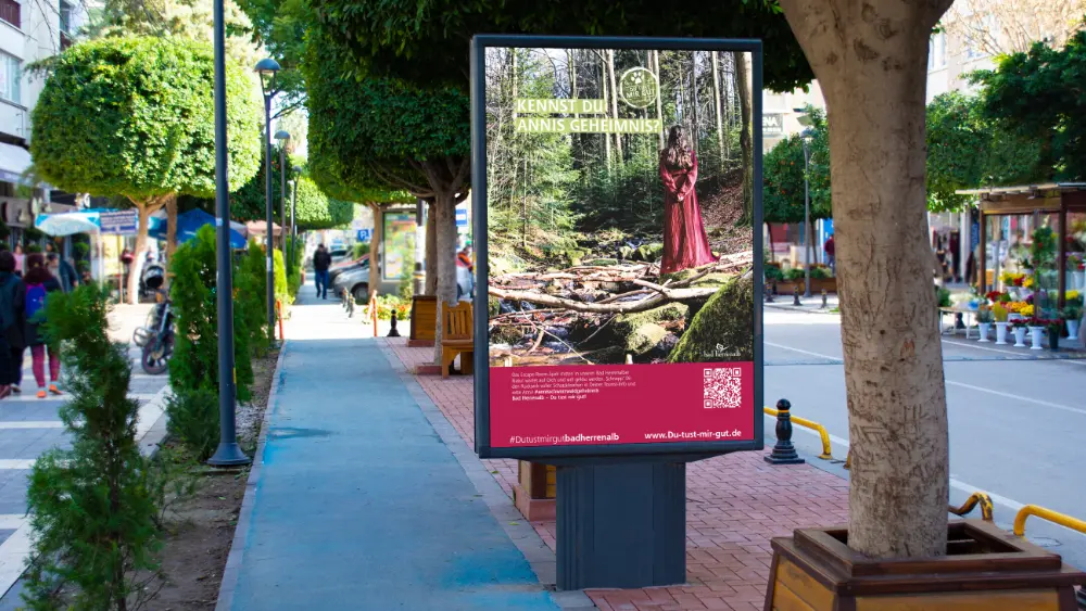 Außenwerbung in einer Stadtstraße für Bad Herrenalb. Das Plakat zeigt eine Frau im roten Kleid im Wald mit der Aufschrift „KENNST DU ANNIS GEHEIMNIS?“. Unten rechts befindet sich ein QR-Code und die Webadresse www.du-tust-mir-gut.de.