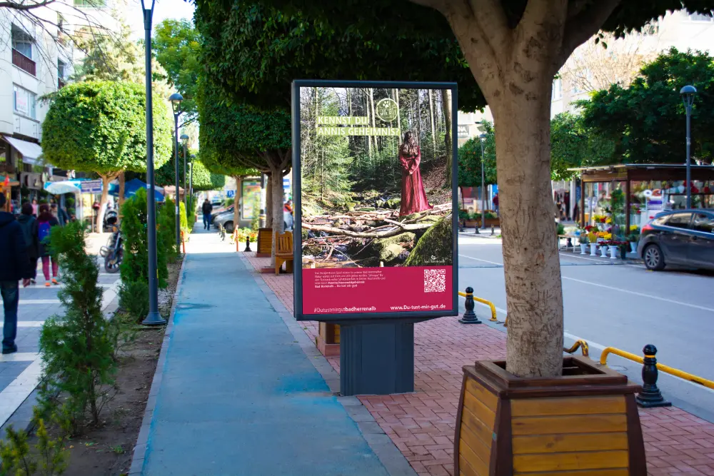 Werbeplakat in einer Stadt mit Waldmotiv und einer Frau im roten Kleid. Text: Kennst du Annis Geheimnis? #Dutustmirgutbadherrenalb, www.Du-tust-mir-gut.de.
