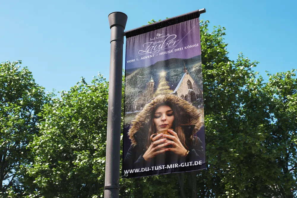 Vertikales Banner an einem Mast: Bad Herrenalb Winterzauber vom 1. Advent bis Heilige Drei Könige. Frau mit Kapuze trinkt aus einer Tasse vor einer Kirche. www.Du-tust-mir-gut.de.