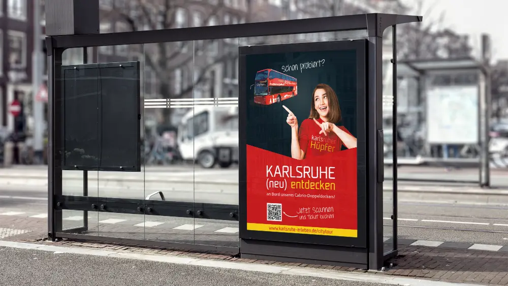 Werbeplakat an einer Bushaltestelle für den „Karlsruher Hüpfer“. Eine lächelnde Frau zeigt auf einen roten Doppeldeckerbus. Text: „Schon probiert? KARLSRUHE (neu) entdecken an Bord unseres Cabrio-Doppeldeckers! Jetzt scannen und Ticket buchen!“. Inklusive QR-Code und URL karlsruhe-erleben.de/citytour.