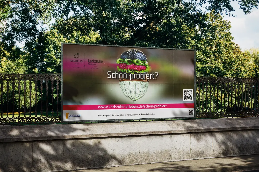 Werbebanner an einem Parkzaun mit einem Burger-Motiv, Michelin-Logo und Text: „Schon probiert?“, „karlsruhe-erleben.de/schon-probiert“ und „railtour“.
