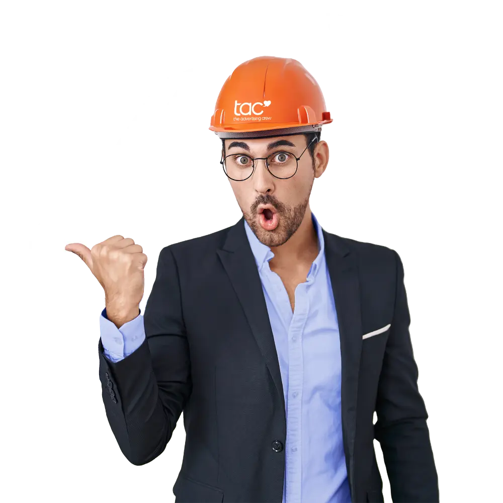 Ein überrascht blickender Mann in Anzug und hellblauem Hemd trägt einen orangefarbenen Schutzhelm mit dem Logo „tac the advertising crew“ und zeigt mit dem Daumen nach links.